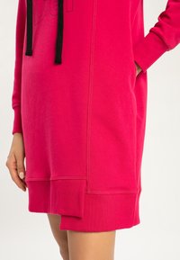 Jasnoróżowa sukienka z kapturem, z czarnymi sznurkami, prążkowanymi mankietami i warstwowym wykończeniem u dołu, z jedną ręką opartą na kieszeni.