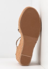 Semelle d'une espadrille compensée marron avec un dessous en caoutchouc texturé et des côtés en jute tressé, positionnée debout contre un fond blanc d'angle.