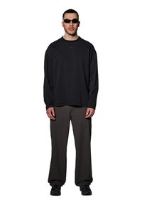 Czarna koszula z długim rękawem o rypsowej strukturze z logo, w połączeniu z luźnymi, ciemnoszarymi spodniami i czarnymi trampkami. Mężczyzna nosi wąskie okulary przeciwsłoneczne.