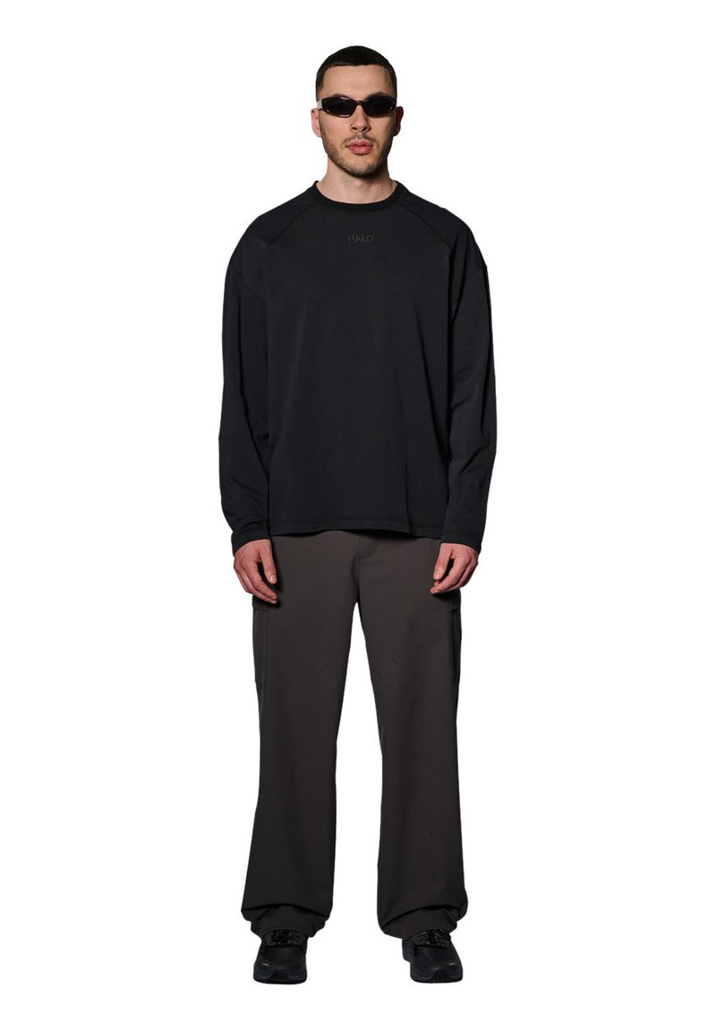 Czarna koszula z długim rękawem o rypsowej strukturze z logo, w połączeniu z luźnymi, ciemnoszarymi spodniami i czarnymi trampkami. Mężczyzna nosi wąskie okulary przeciwsłoneczne.