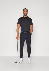 Black polo shirt with short sleeves and a collar, paired with dark trousers. Accessories include a black belt and white sneakers. Holding a golf club.