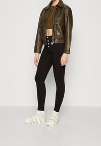Chaqueta corta de cuero marrón, top oliva, vaqueros skinny de tiro alto negros con cierre de botón, y zapatillas deportivas blancas con cintas.
