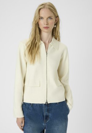 Veste zippée de couleur crème avec un col, deux poches avant et une coupe ajustée. Portée par-dessus un jean bleu légèrement usé.