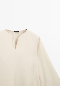Blouse beige clair avec un col en V, des manches longues, une texture lisse et des accents de couture subtils. Design simple et minimaliste.