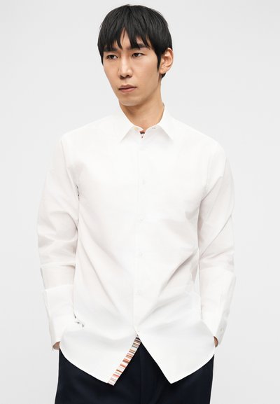 Άνδρας με κοντά μαύρα μαλλιά, ντυμένος με λευκό πουκάμισο με κουμπιά και μαύρο παντελόνι, στέκεται με τα χέρια στις τσέπες μπροστά σε απλό λευκό φόντο.