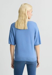 Tricou din lână albastră cu o croială lejeră, mâneci scurte și tiv cu nervuri. Țesătura are o textură fină și un luciu subtil.