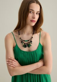 Collana dichiarativa multicolore con frange verdi e accenti neri, caratterizzata da perle testurizzate e dettagli in metallo dorato, indossata sopra un vestito verde.