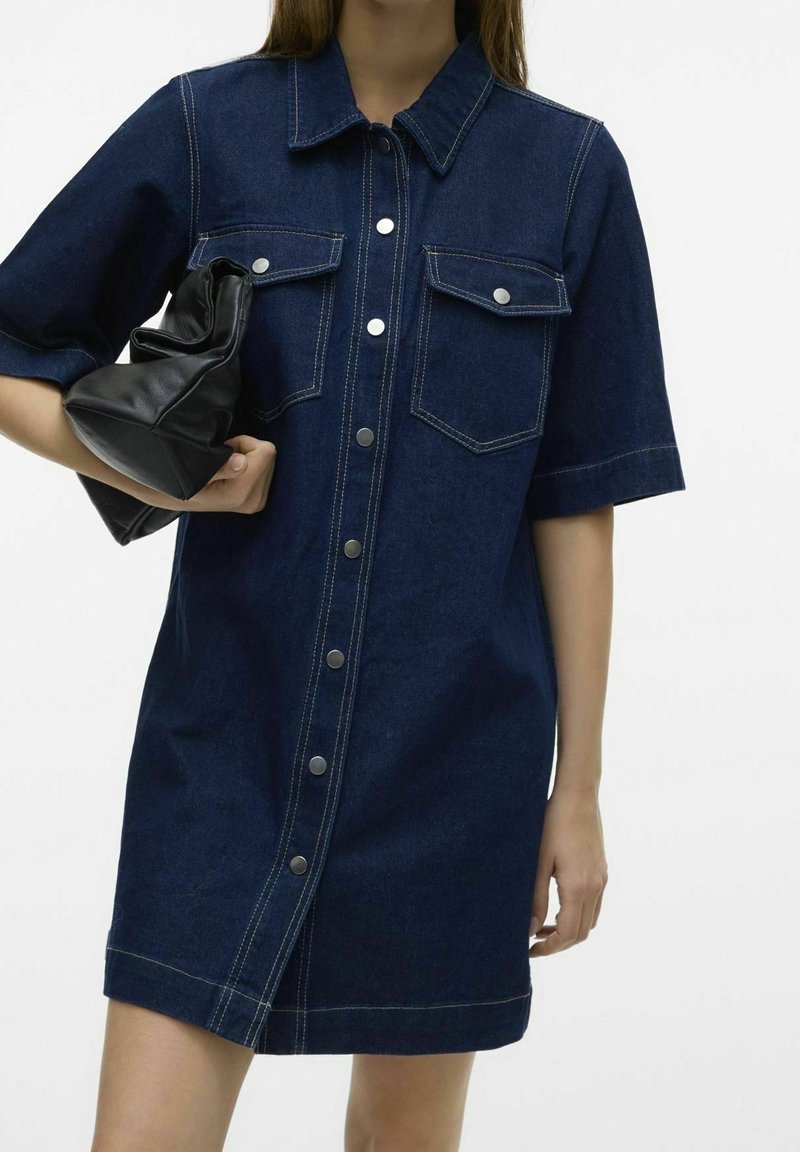 Femme portant une robe en jean bleu foncé à manches courtes, boutonnée sur le devant avec des poches poitrine, tenant une pochette en cuir noire sous le bras.