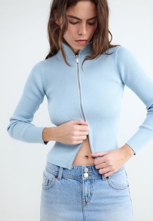 Kvinde iført lyseblå ribstrikket trøje med lynlås, delvist åbnet for at afsløre hendes mave, kombineret med højttaljede lyse denim jeans.
