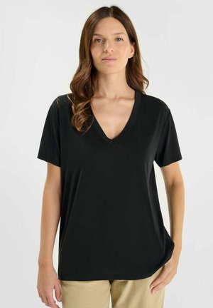 Vrouw met lang bruin haar, gekleed in een zwart V-hals T-shirt met korte mouwen en beige broek, staande tegen een effen lichte achtergrond.