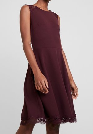 Burgundy ärmlös klänning med en figurnära överdel, utsvängd kjol, spetsdetaljer på axlarna och en scalloped spetskant. Slät textil.