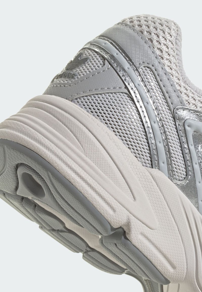 Zapatilla deportiva gris con una parte superior de malla transpirable y acentos plateados, suela de goma gruesa y detalles texturizados en la suela exterior.