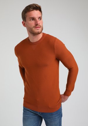 Ribbed long-sleeve sweater in rust orange, featuring a round neckline, fitted silhouette, and textured fabric. Paired with denim jeans.