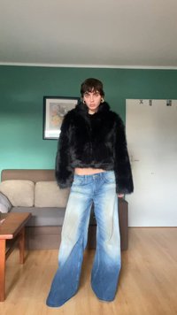 Fekete műszőrme cropped dzseki, széles lábú, hanyatló kék farmerruha világoskék gradienstel, fekete felsővel viselve, zöld háttér előtt.