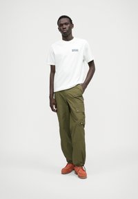 T-shirt blanche à manches courtes avec un graphisme bleu sur la poitrine, associée à un pantalon cargo ample vert olive et des chaussures brunes à lacets.