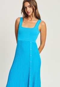 A sleeveless, bright blue dress featuring a square neckline, fitted bodice, and a pleated skirt with contrasting buttons down the front.
