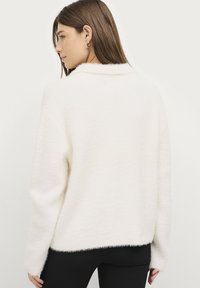 Witte, fluffy trui met een licht oversized, relaxte pasvorm, voorzien van een brede kraag en lange mouwen, gecombineerd met zwarte broek.