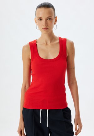 Top sin mangas de canalé rojo con un amplio escote redondo, combinado con pantalones oscuros que tienen cordones blancos.