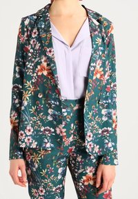 Floral print suit in teal fabric, featuring a blazer with a lapel and matching trousers. Blouse in light lavender underneath.