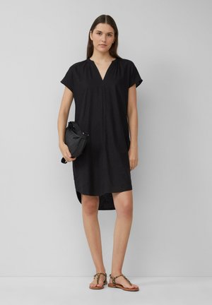 Schwarzes Leinenkleid mit kurzen Ärmeln und V-Ausschnitt. Verfügt über einen seitlichen Schlitzsaum und wird mit flachen, braunen Sandalen mit Kettendetails kombiniert.