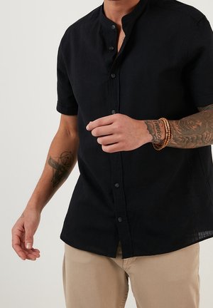 Man met een zwart, kort mouwig, knoopjeshemd met tatoeages op beide armen en een kralenarmband om de rechterpols, beige broek.