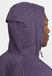Hombre con una chaqueta con capucha texturizada de color púrpura, mostrado desde atrás y de perfil lateral con la capucha completamente puesta.