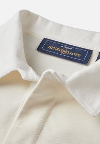 Polo shirt color crema con tessuto a trama, caratterizzato da un colletto ricamato e un'etichetta blu navy con scritta dorata "HENRI LLOYD."
