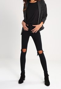 Femme debout, mains sur les hanches, portant un jean skinny noir déchiré, un haut noir, un cardigan gris foncé ouvert devant, et des bottines noires.