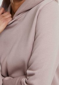 Sudadera de color rosa claro con una tela suave y texturizada. Presenta una gran capucha caída y un ajuste relajado, con detalles de costura visibles.