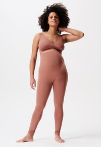 Zwangere vrouw met krullend haar die blootsvoets staat in een ribbelige, bijpassende, stoffige rozenkleurige sportoutfit tegen een vlakke witte achtergrond.