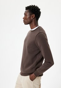 Sudadera marrón con escote redondo, puños y dobladillo acanalados. La tela parece suave con una textura lisa, usada sobre pantalones de color claro.