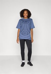 Blue oversized t-shirt with a graphic of the Eiffel Tower and "PARIS SAINT-GERMAIN" text, paired with black fitted pants and black footwear.