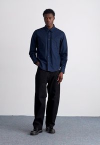 Camicia blu navy a maniche lunghe con colletto strutturato, due tasche sul petto, abbinata a pantaloni neri a gamba larga e scarpe nere.