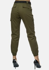 Pantalons cargo en vert olive, dotés de poignets et d'une taille élastiques, avec plusieurs poches à l'arrière et sur les côtés. Portés avec des talons noirs.