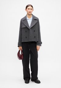Šedý vlněný kabát typu pea coat s černými dvojitými knoflíky, pod ním světle modrá pruhovaná košile, černé kalhoty širokého střihu a bordó kabelka.