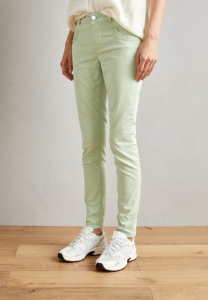 Personne portant un pantalon slim vert clair et des baskets blanches, debout sur un sol en bois devant un mur uni.
