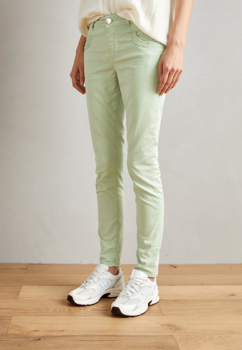Personne portant un pantalon slim vert clair et des baskets blanches, debout sur un sol en bois devant un mur uni.