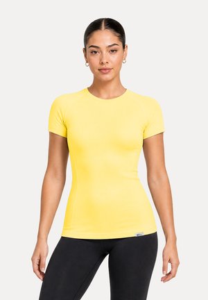 Femme aux cheveux foncés attachés en chignon, portant de petites boucles d’oreilles en créoles, un haut jaune cintré à manches courtes et des leggings noirs sur un fond uni.