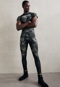 Athletic man with crossed arms wearing black and gold graphic compression leggings and matching short-sleeve top standing against a plain wall.