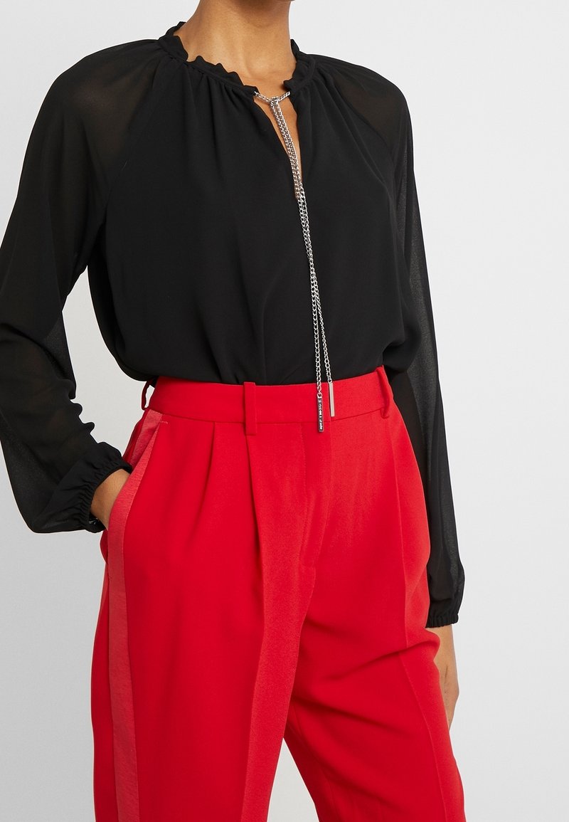 Blouse noire à manches longues avec un col froufrouté, rentrée dans un pantalon rouge taille haute avec poches latérales, associée à un collier chaîne argenté.