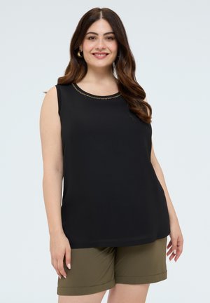 Woman with long wavy hair wears a black sleeveless top with subtle neckline detail and olive green shorts, smiling against a light background.