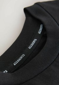 Primer plano de una prenda negra con cuello redondo que muestra la etiqueta interior cosida con el nombre de la marca "ALLSAINTS" en texto blanco.