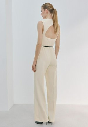 Femme avec une queue de cheval blonde portant une combinaison beige sans manches à dos ouvert et une ceinture noire étroite, debout devant un fond blanc uni.