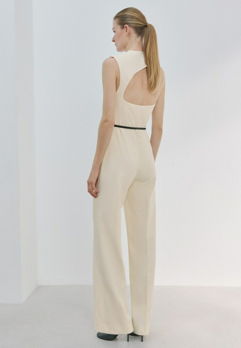 Žena s blond culíkem, která má na sobě béžový overal bez rukávů s odhalenými zády a úzkým černým pásem, stojí před jednoduchým bílým pozadím.