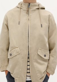Giacca beige con cappuccio realizzata in materiale morbido, dotata di zip anteriore, due ampie tasche a soffietto e una finitura testurizzata. Camicia a righe sotto.