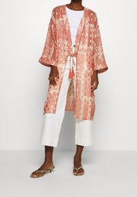 Lehký kimono s vzorem v broskvové a bílé barvě, se zavazováním na tkaničku a střapcem. Má široké rukávy a otevřený přední střih.