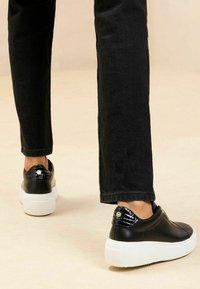 Sneakers in pelle nera con tallone texturizzato in pelle di coccodrillo nera, suola bianca e dettaglio metallico sul retro. Indossati con jeans neri.
