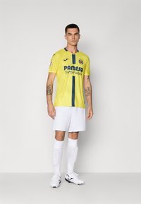 Mees seisab kollases Villarreal CF jalgpallivormis, valgetes šortsides, sokkides ja tossudes, lihtsa heleda tausta taustal.