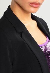 Blazer noir avec des revers nets, un tissu lisse et une coupe ajustée sur un haut violet à paillettes, mettant en avant une coupe moderne.