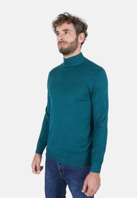 Maglione dolcevita turchese realizzato in tessuto morbido, maniche lunghe, design aderente, polsini e orlo a coste, abbinato a jeans scuri.